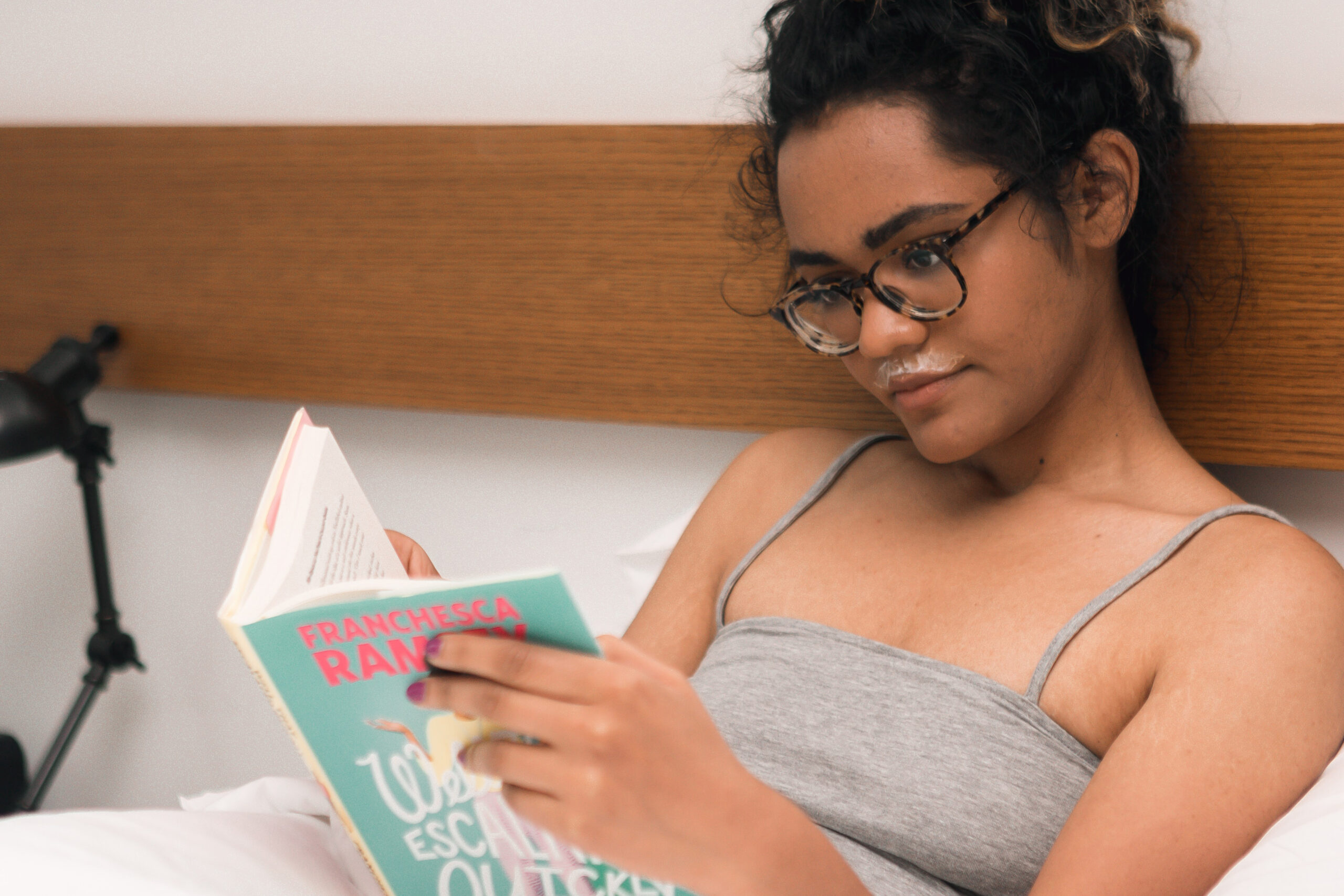 Young femme in grey tank and dark-rimmed eyeglasses in bed reading a brightly colored book.