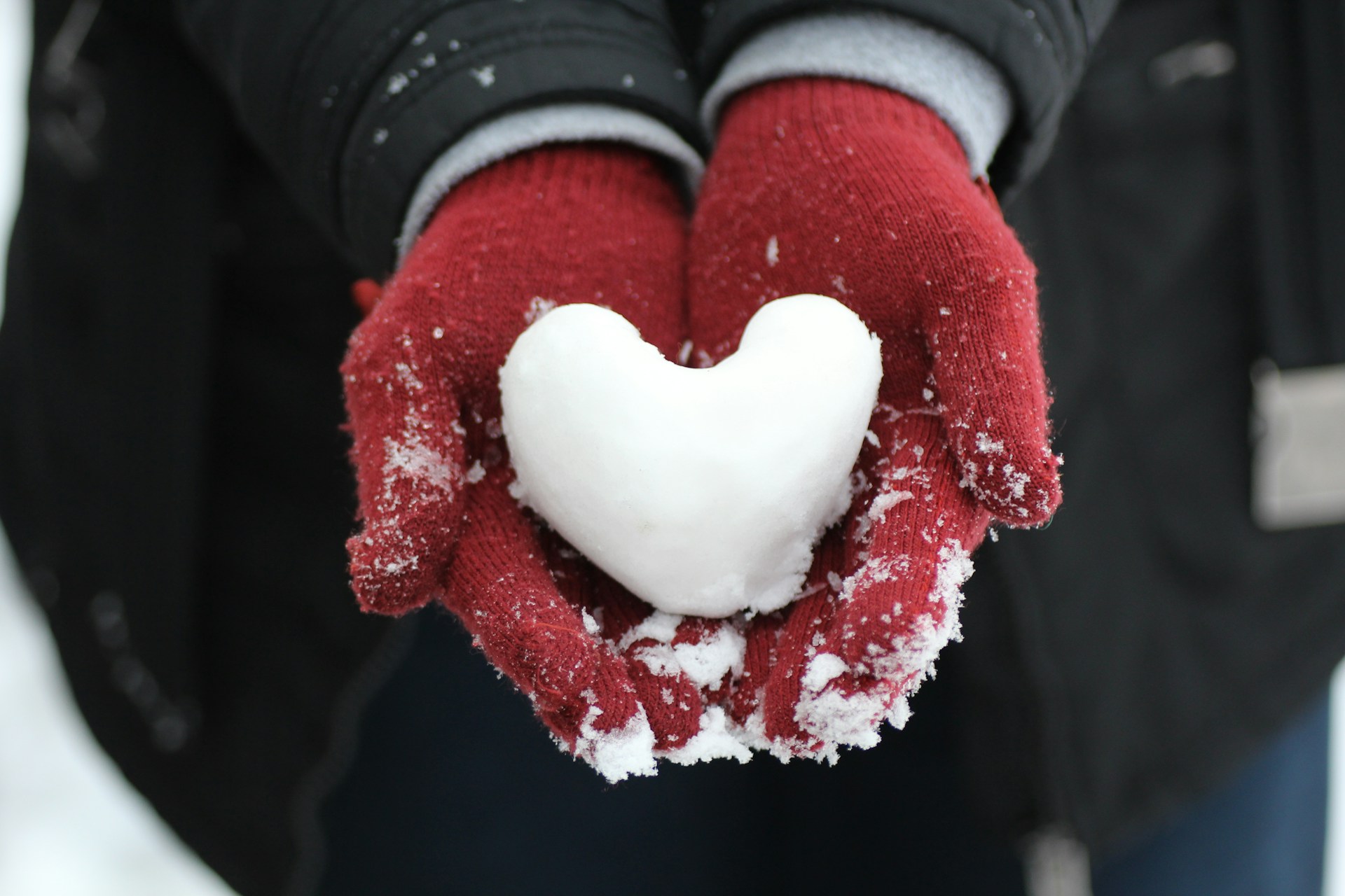 A person wearing red gloves holds a heart-shaped snowball